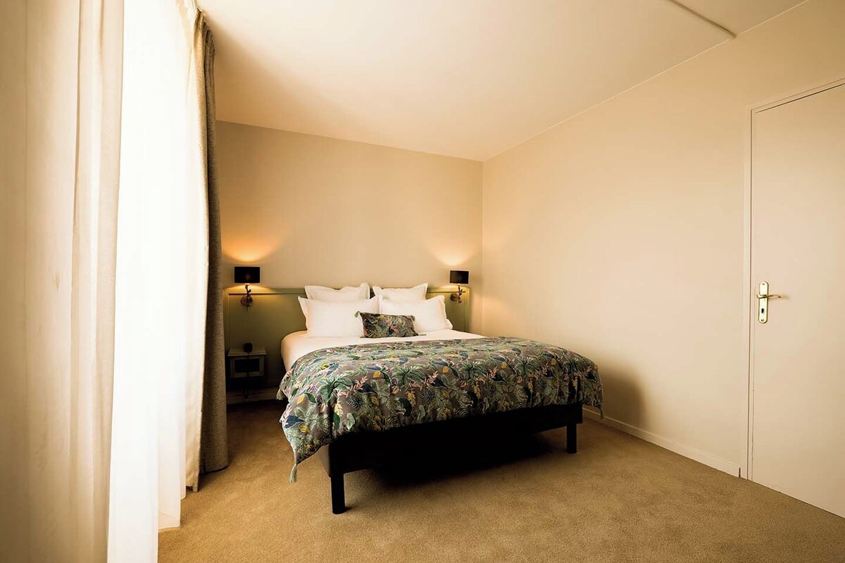 Un dormitorio con cama, dos mesitas de noche, una mesita auxiliar, una puerta y una cortina.
