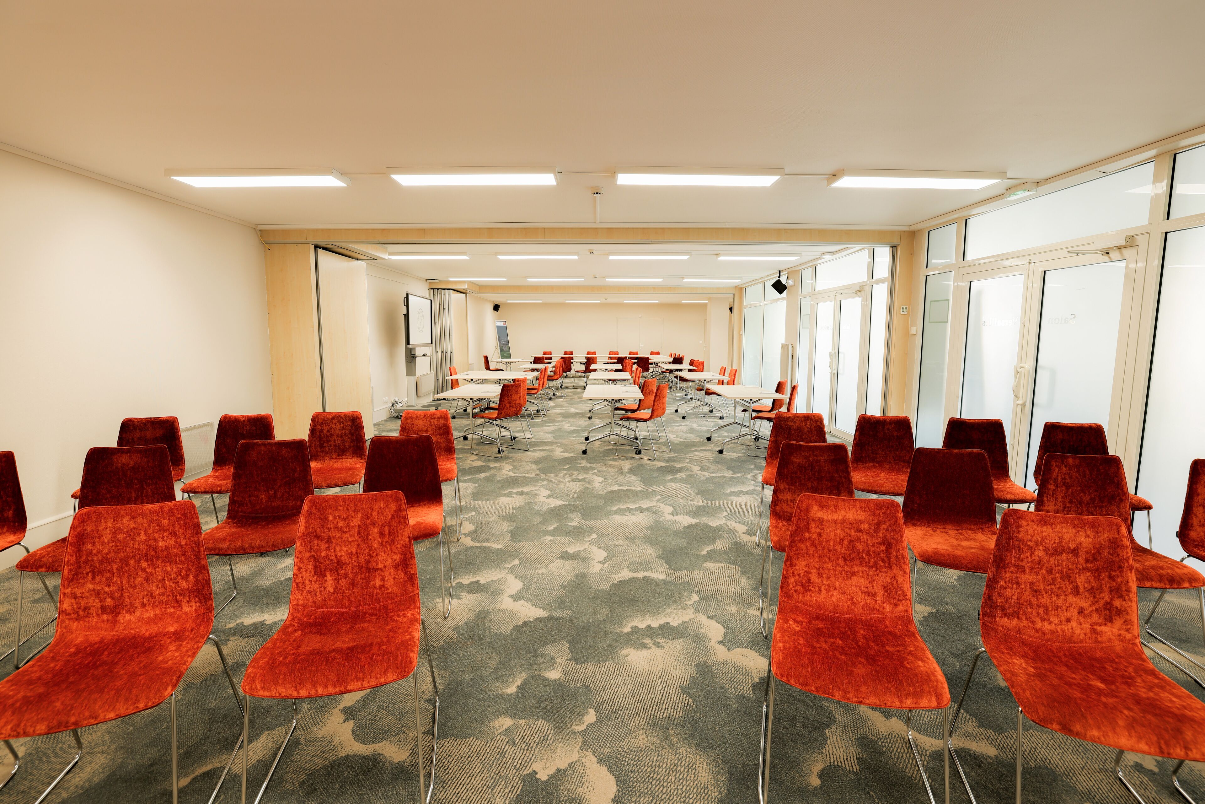 A conference room with rows of orange chairs, a carpeted floor, and large windows on one side.