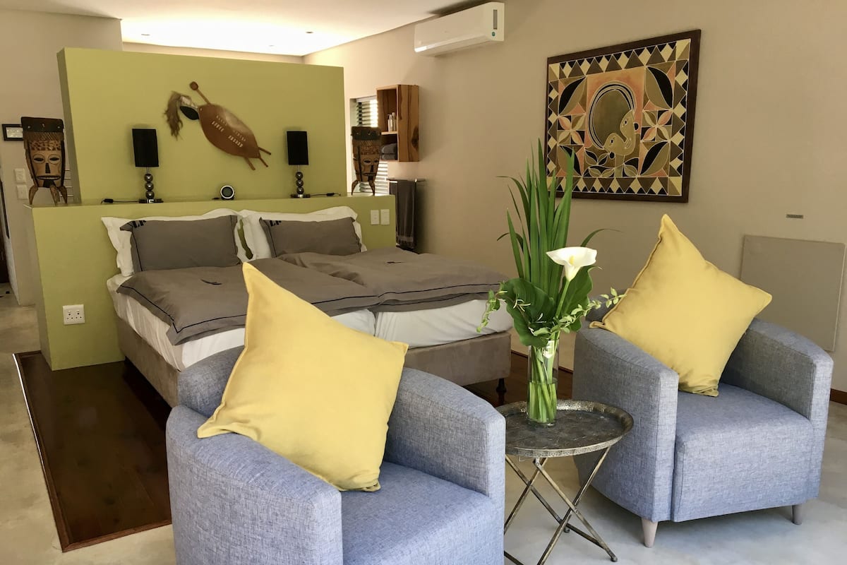 A modern bedroom with a bed, two armchairs, a small table, and a decorative painting.
