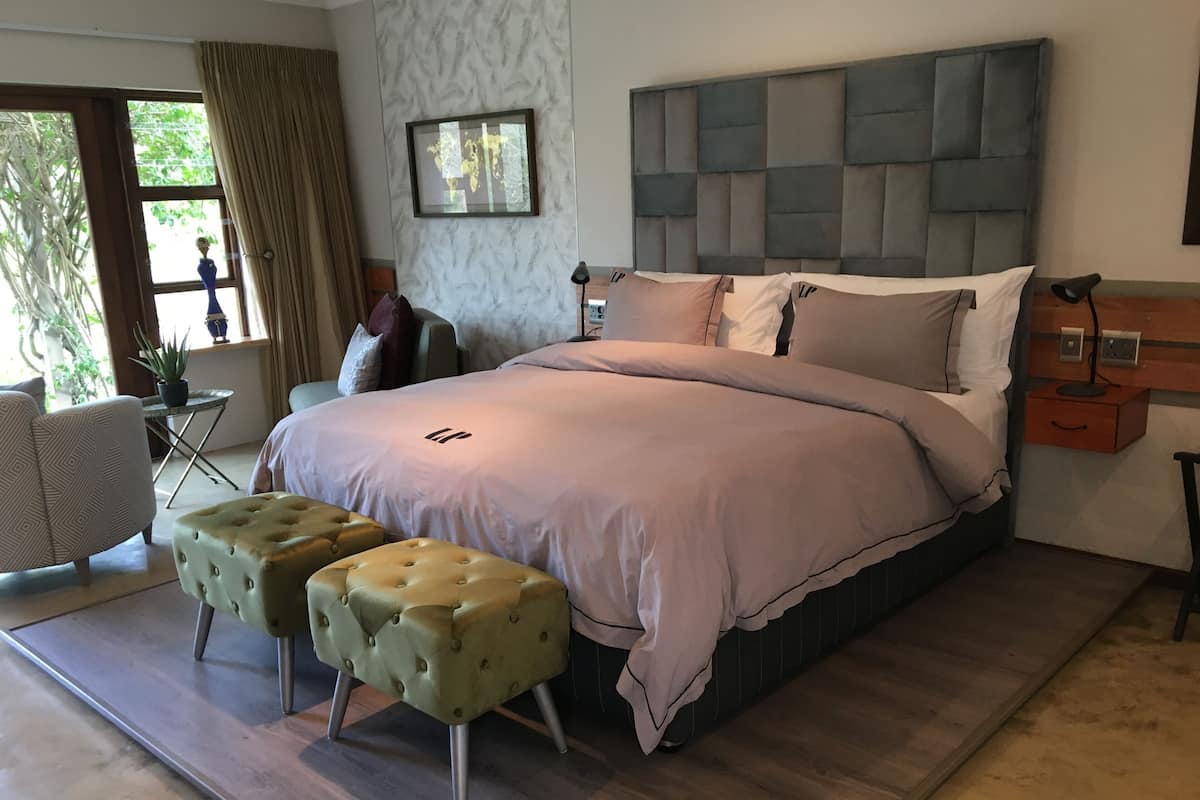 A modern bedroom with a large bed, two tufted ottomans, a headboard with a world map, and a small table with a lamp.