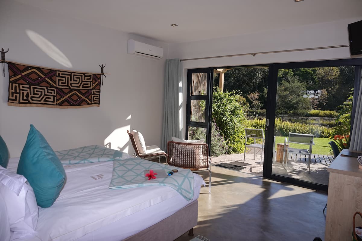 A bedroom with a large bed, a view of the garden through a sliding door, and a small seating area with chairs.