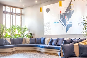 Un sofá curvo con cojines azules, una obra de arte geométrica en la pared y una ventana con persianas.