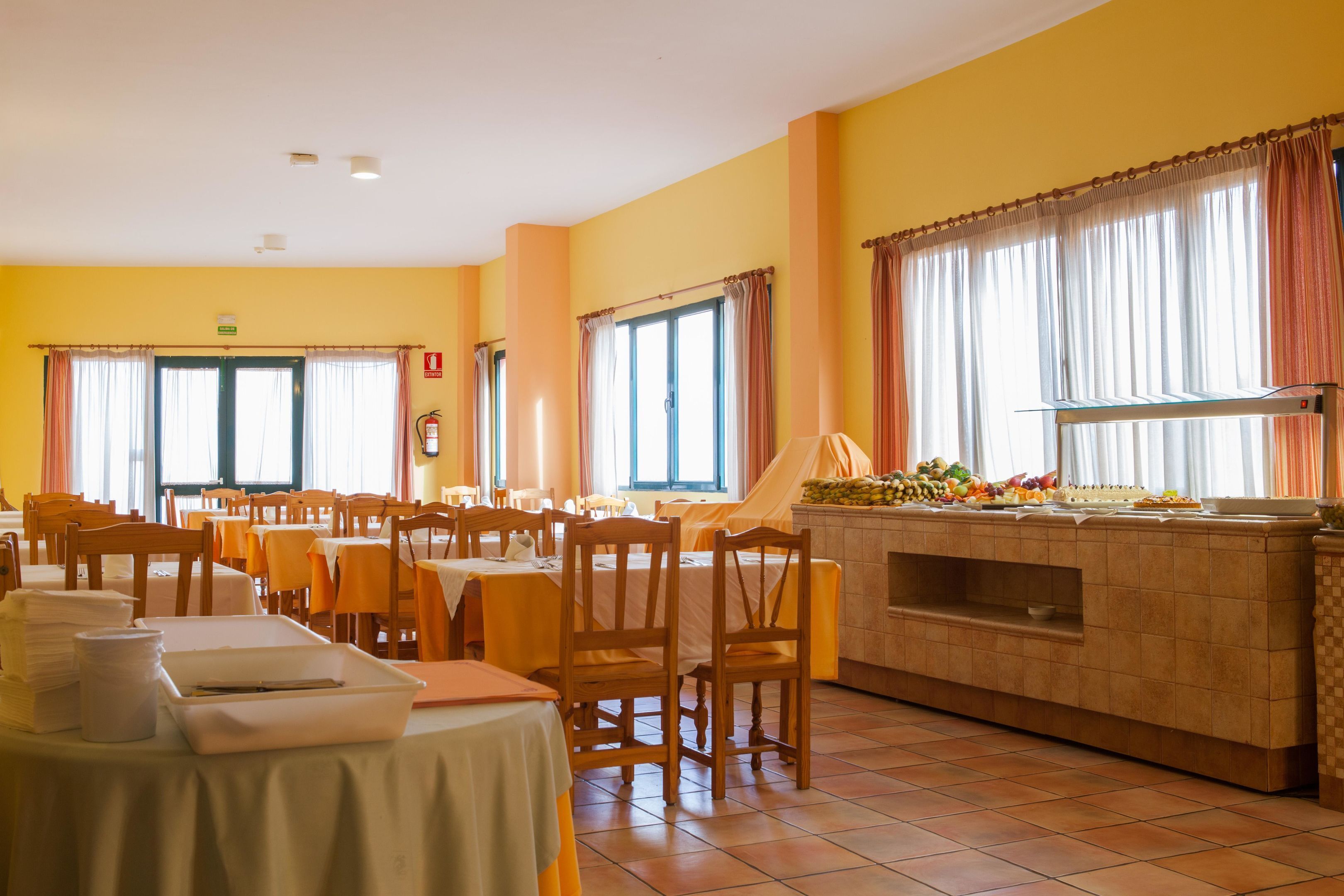 Een eetruimte met meerdere tafels en stoelen, een buffet met eten, en grote ramen voorzien van gordijnen.
