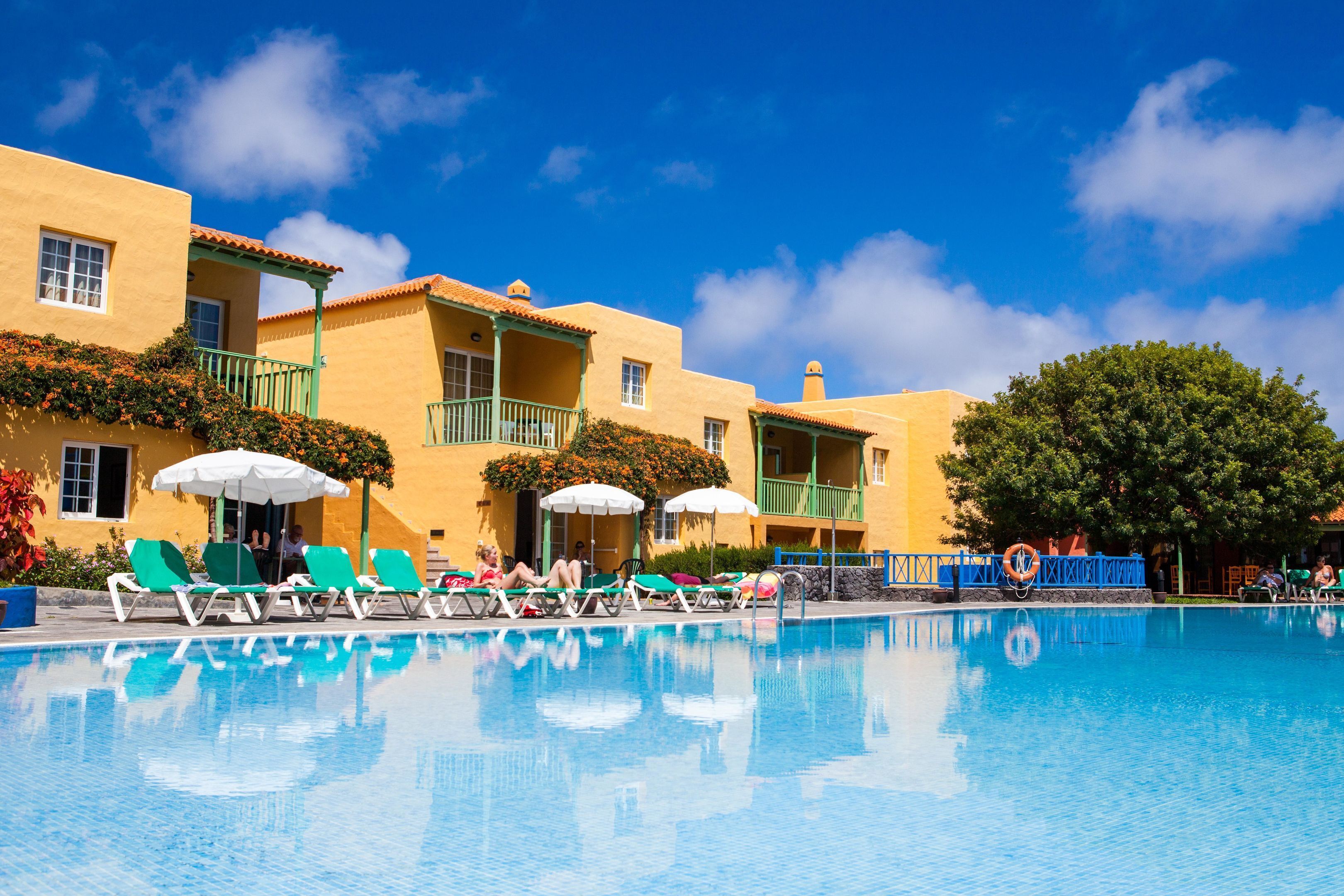 A pool area with lounge chairs and umbrellas, a clear blue sky, and a building with balconies.