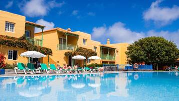 A pool area with lounge chairs and umbrellas, a clear blue sky, and a building with balconies.