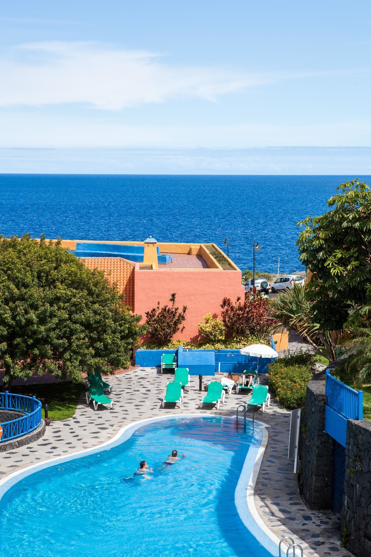 A swimming pool with two people swimming, surrounded by lounge chairs and a view of the ocean.