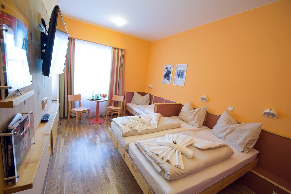 A hotel room with a wooden floor, two beds, a desk with a chair, a window with curtains, and a wall-mounted TV.