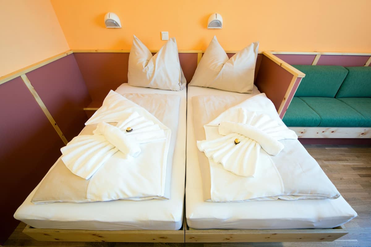 A room with two beds, each with white linens and pillows, a green sofa, and a wall-mounted light fixture.
