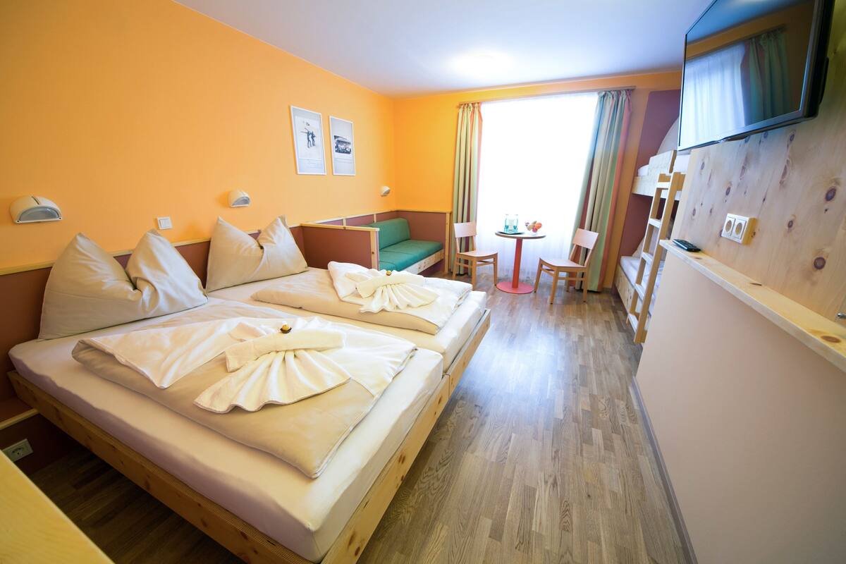 A hotel room with a double bed, a small sofa, a TV, a table with a chair, and a window with curtains.