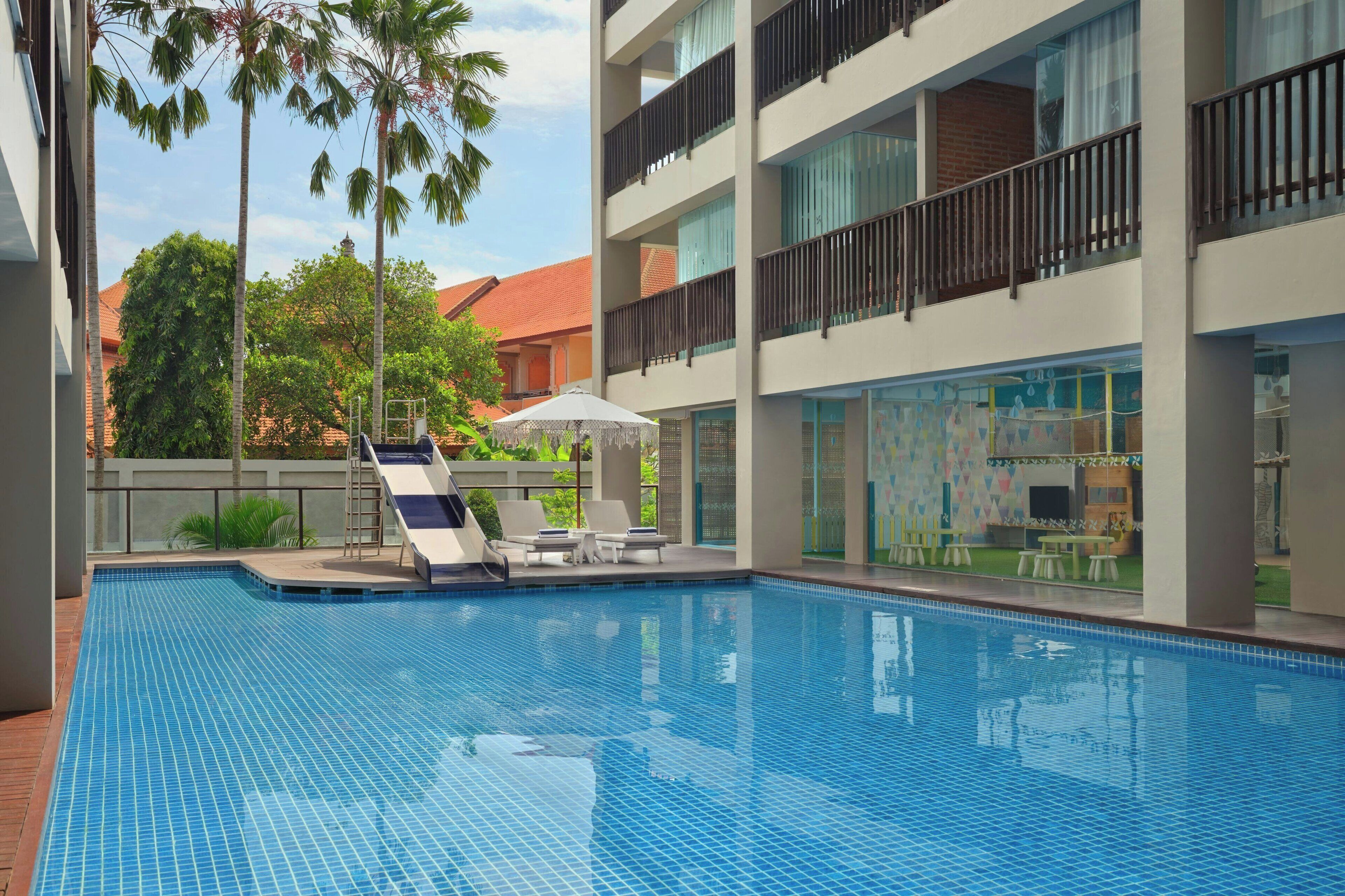Una piscina con sillas de descanso y sombrillas, rodeada por un edificio moderno con balcones y ventanas amplias.