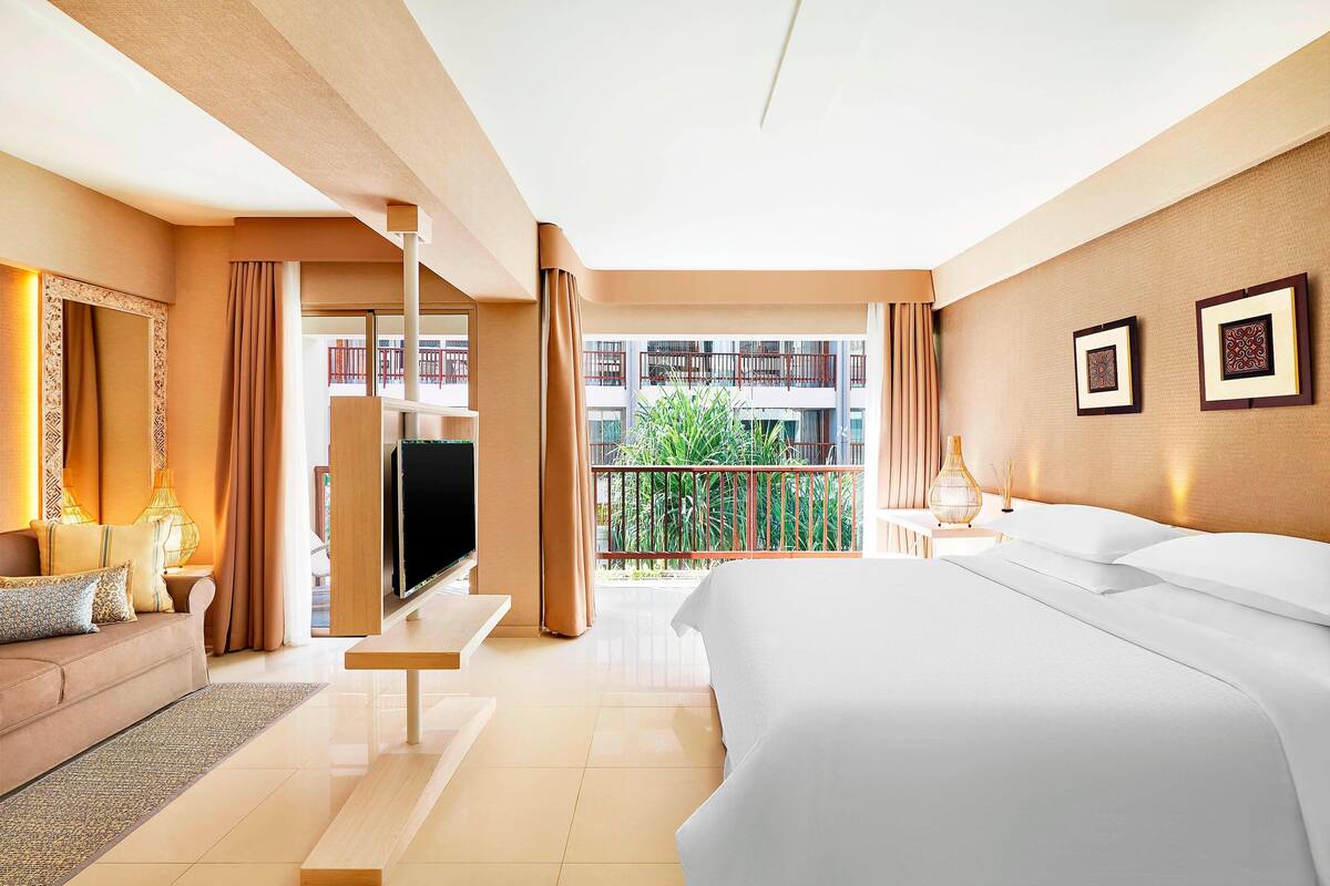 A hotel room with a bed, a TV, a sofa, and a balcony with a view of a building and greenery.