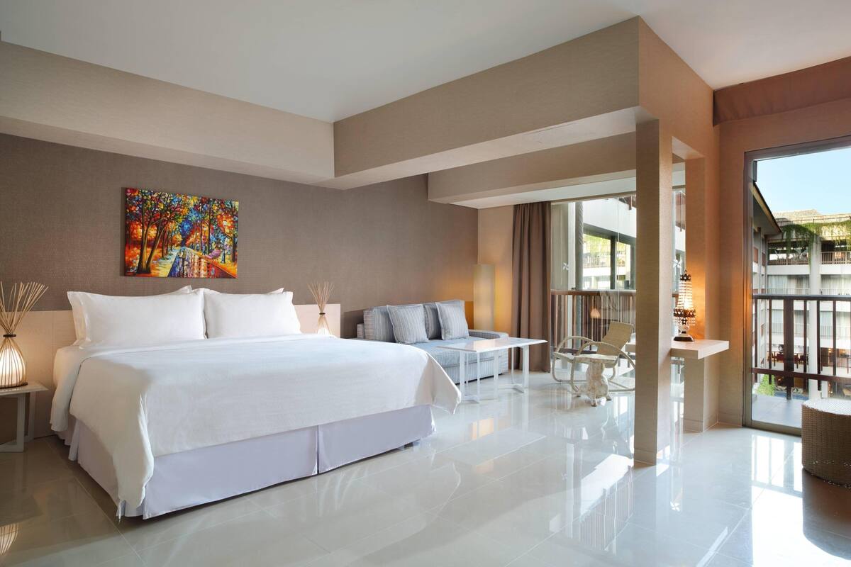 A modern hotel room with a large bed, a sofa, a dining area, and a balcony with a view.