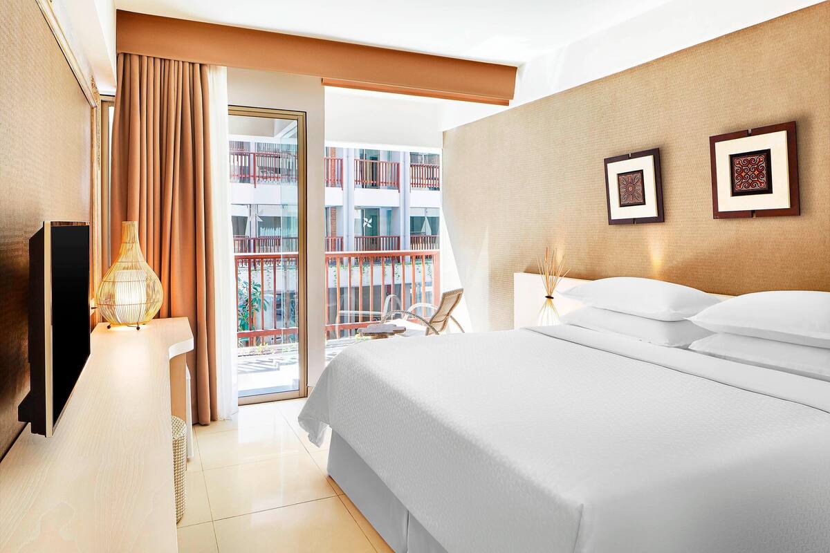 A hotel room with a large bed, a TV, a lamp, and a view of a balcony with a railing.