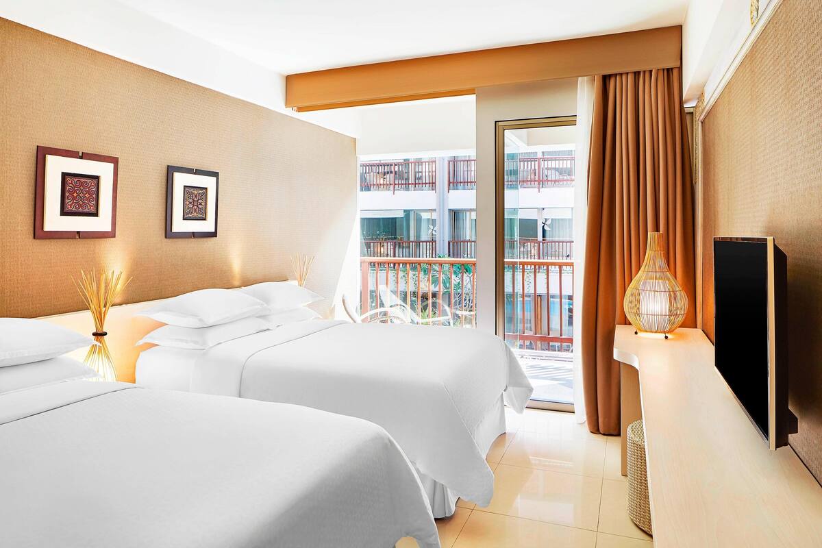 A hotel room with two beds, a TV, a balcony with a view of a building, and a large window with curtains.