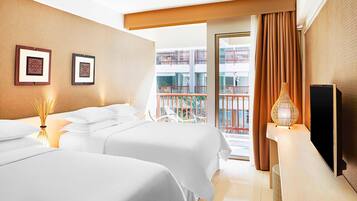 Une chambre d’hôtel avec deux lits, une télévision, un balcon donnant sur un immeuble et une grande fenêtre avec des rideaux.