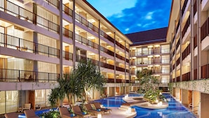 A multi-story hotel with a swimming pool, sun loungers, and a well-lit interior. - Four Points By Sheraton Bali, Kuta (Kuta)