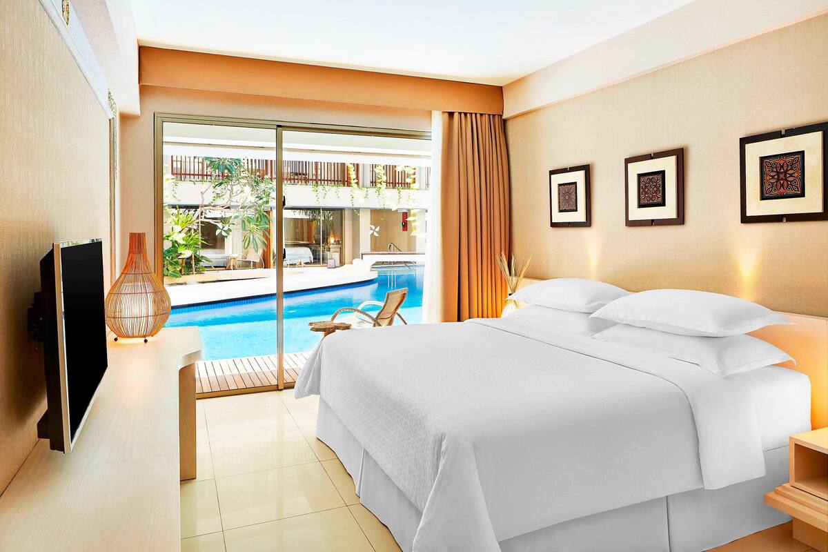 A hotel room with a bed, a TV, a bedside table, a lamp, and a view of a pool and outdoor seating area.