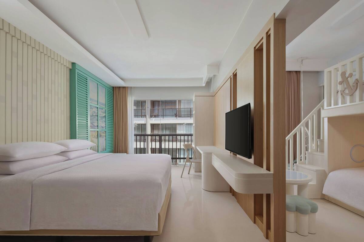 A modern hotel room with a large bed, a desk with a flat-screen TV, a small table, and a balcony with a railing.