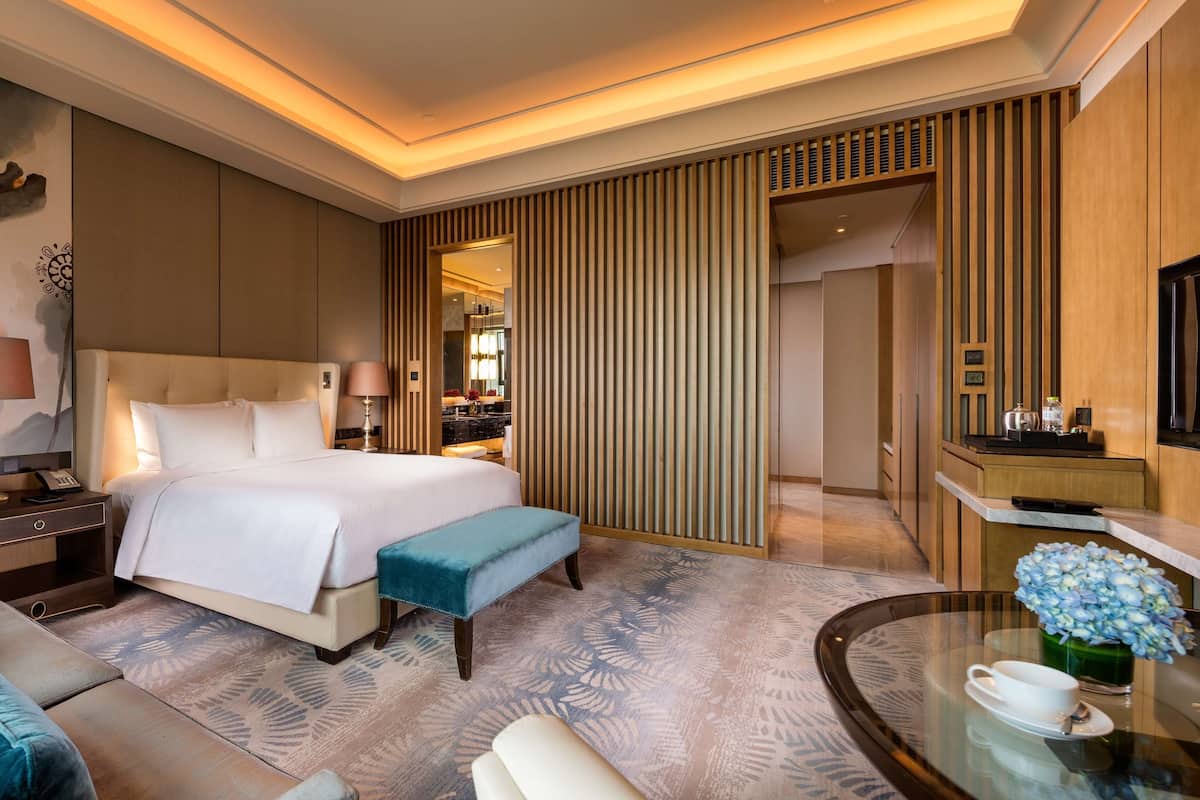 Una habitación de hotel moderna con una cama grande, una zona de estar con sofá y sillón, una mesita con una taza y flores, y un televisor en la pared.