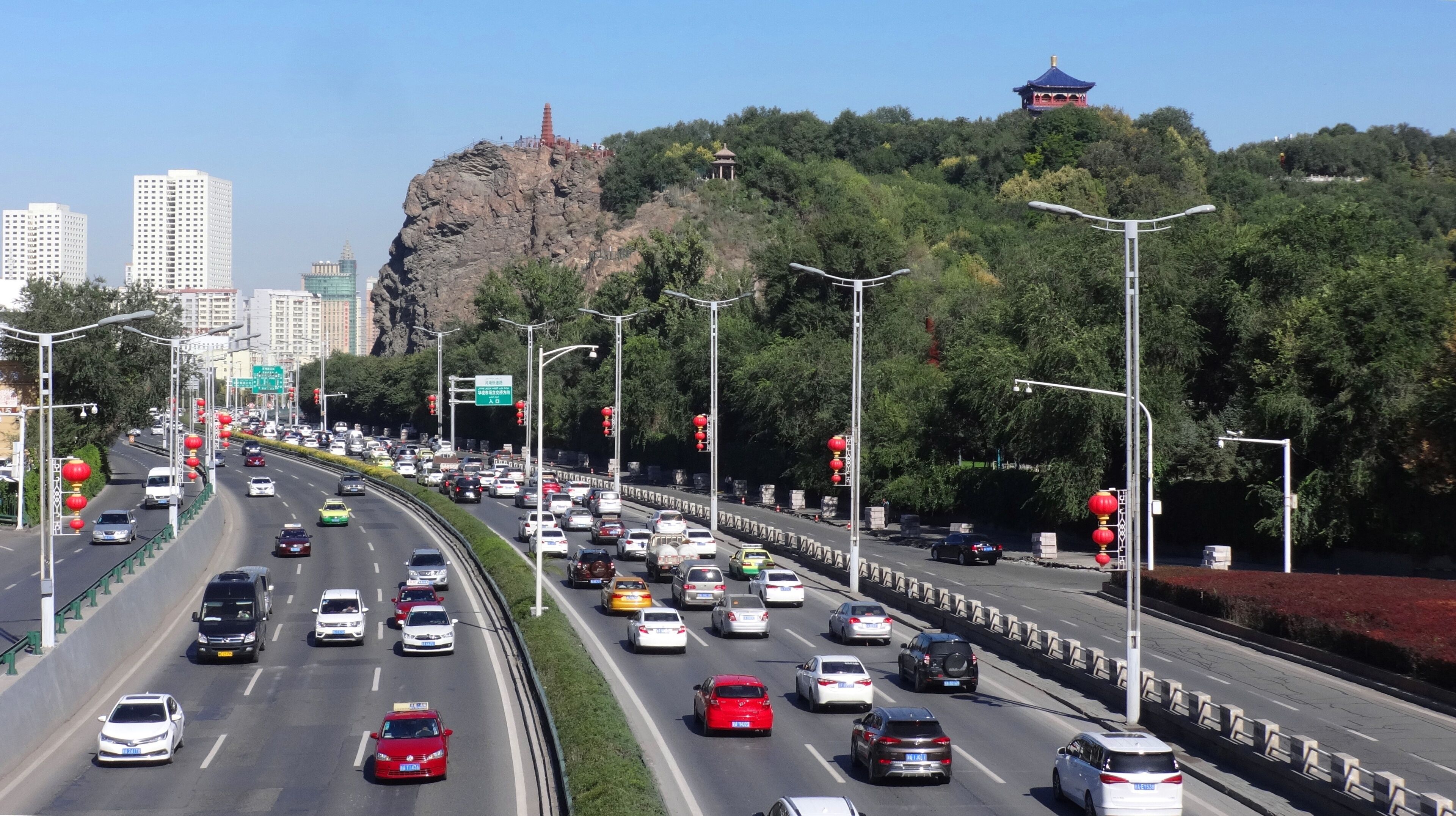 Una autopista con varios carriles y vehículos, rodeada de zonas verdes y edificios urbanos.