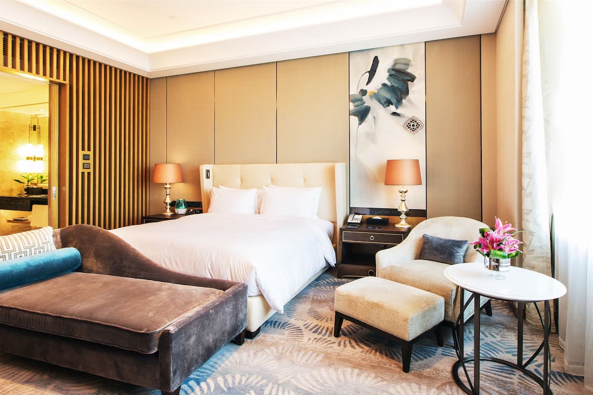 Habitación de hotel con una cama grande, dos mesitas de noche con lámparas, un sofá, un reposapiés, una mesita con un jarrón de flores y un cuadro en la pared.