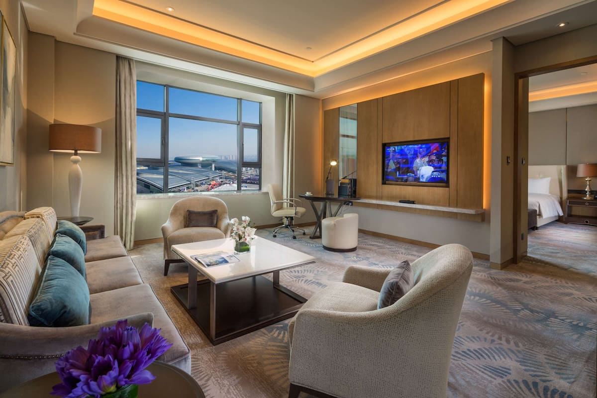 Una habitación de hotel moderna con un ventanal grande, un televisor de pantalla plana, un sofá, una mesa baja y una mesa de comedor.