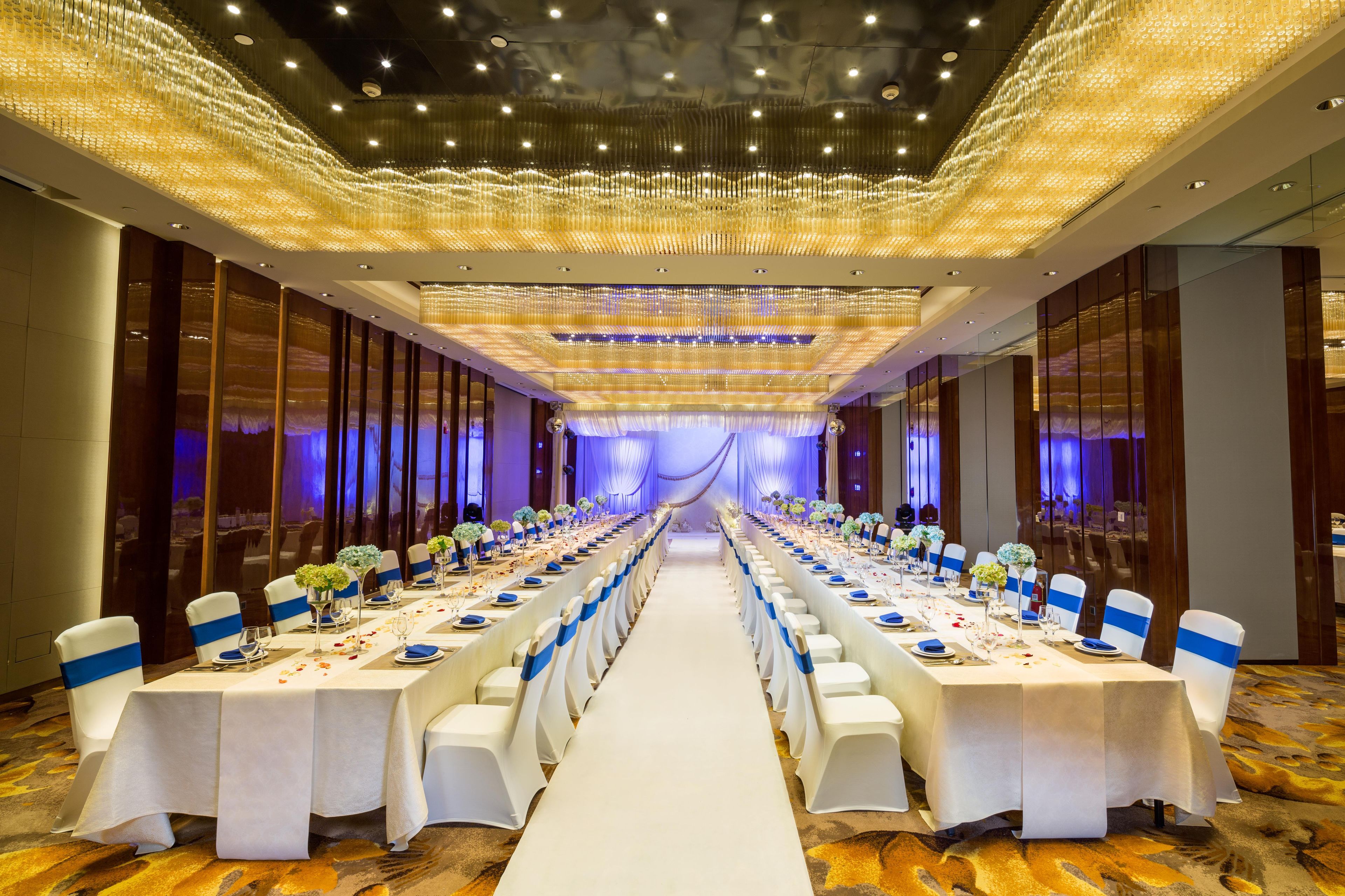 Une salle de banquet avec de longues tables, des nappes blanches et des rubans bleus sur les chaises.