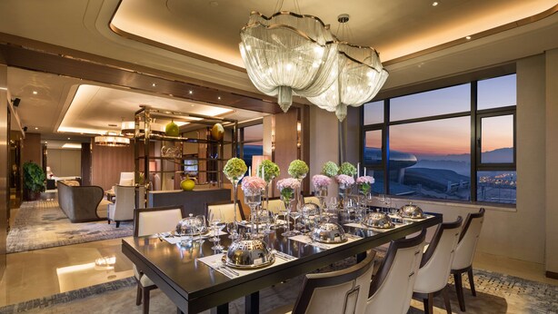 A modern dining area with a large table set for six, surrounded by chairs. A chandelier hangs above, and the room features large windows with a view of the cityscape. - Hilton Urumqi (Ürümqi)