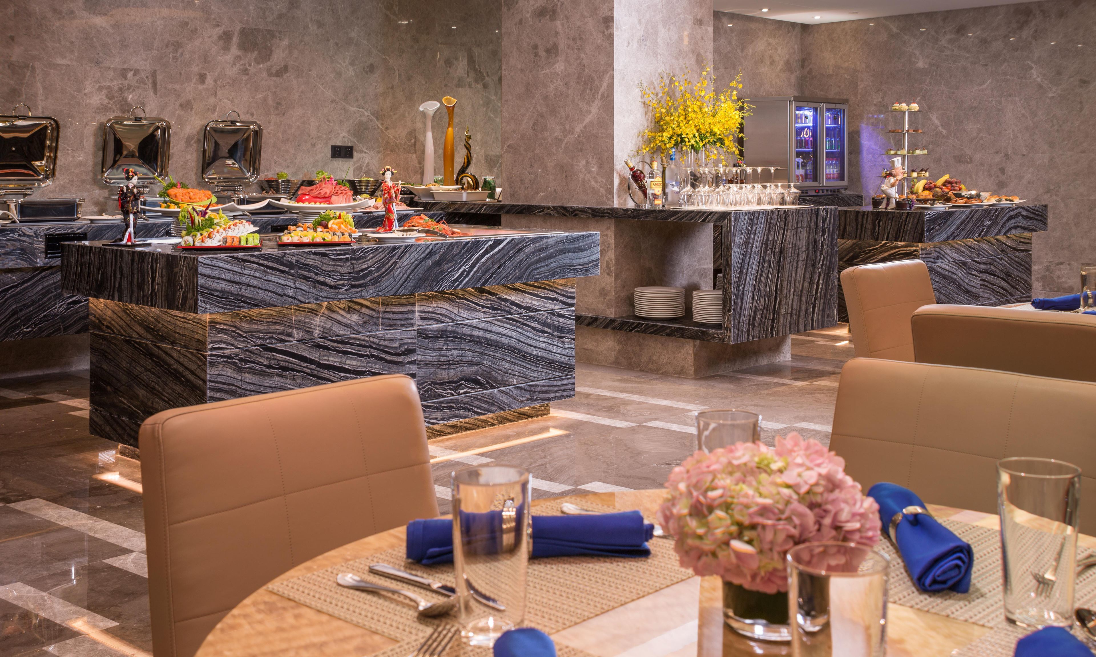 A hotel dining area with marble countertops, a buffet with various food items, and a bar area with a refrigerator.