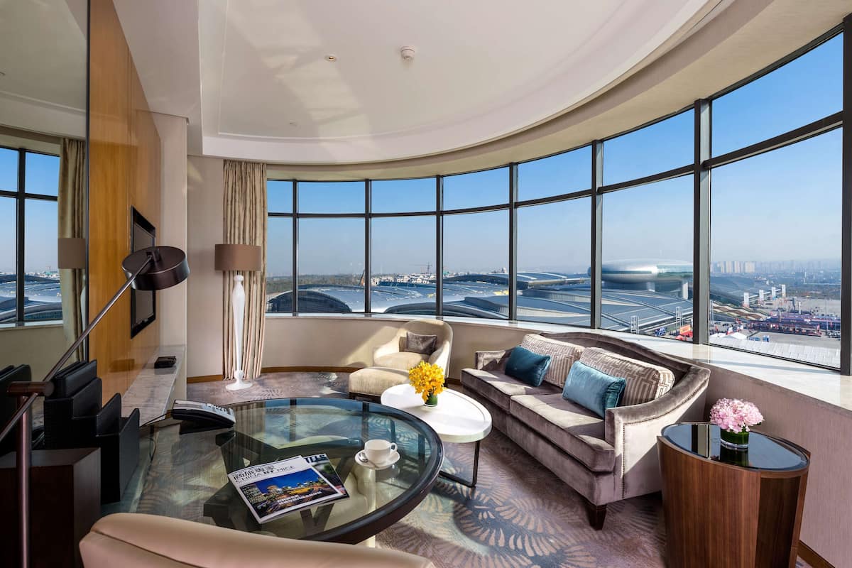 Una habitación de hotel moderna con techo curvo, ventanas panorámicas amplias, una mesa de centro de vidrio, un sofá y una pequeña mesita de noche con flores.