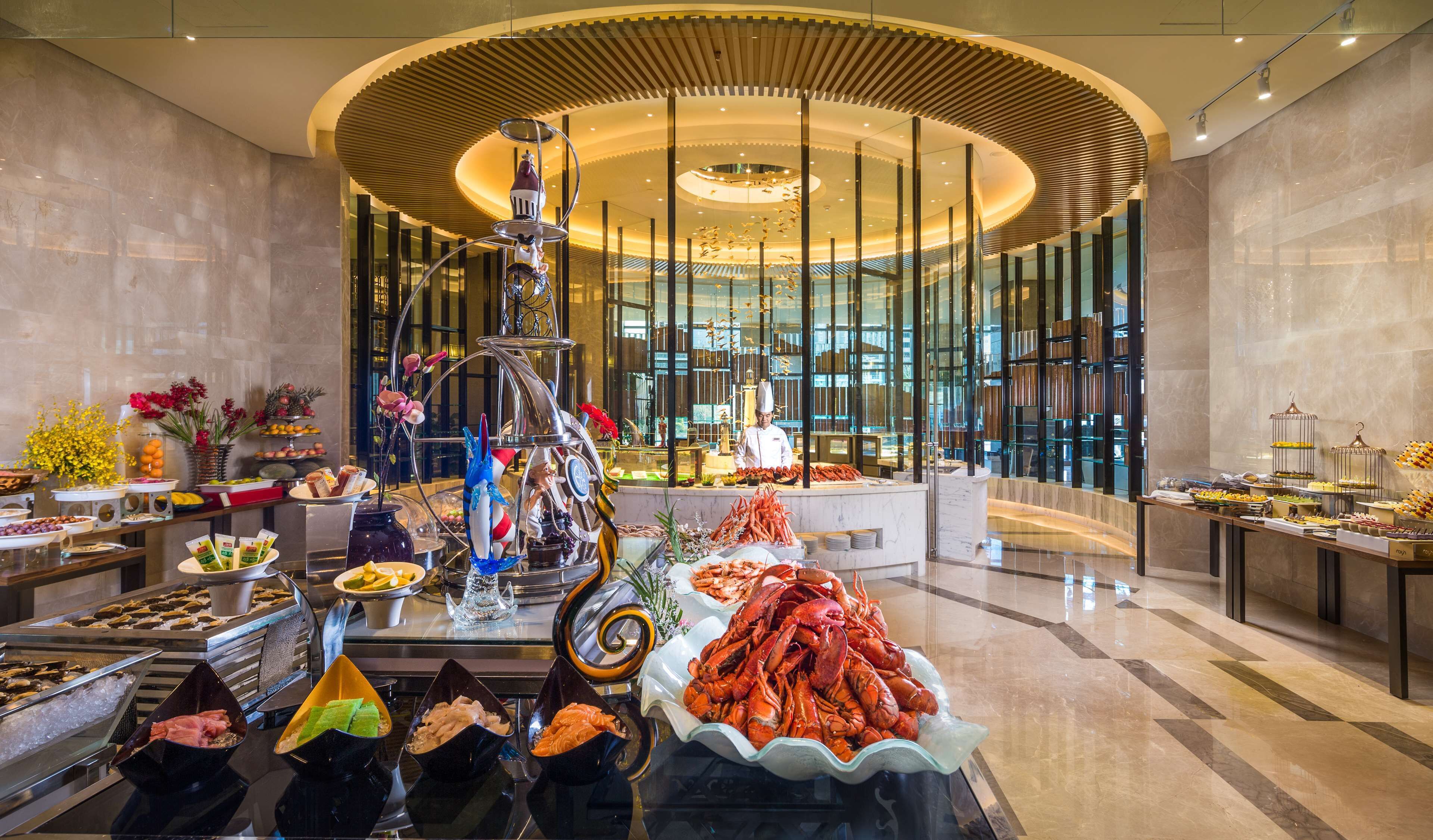 Un lujoso bufé con mariscos, frutas y pastelería, ubicado en un moderno vestíbulo de hotel con un amplio ventanal y una fuente decorativa.