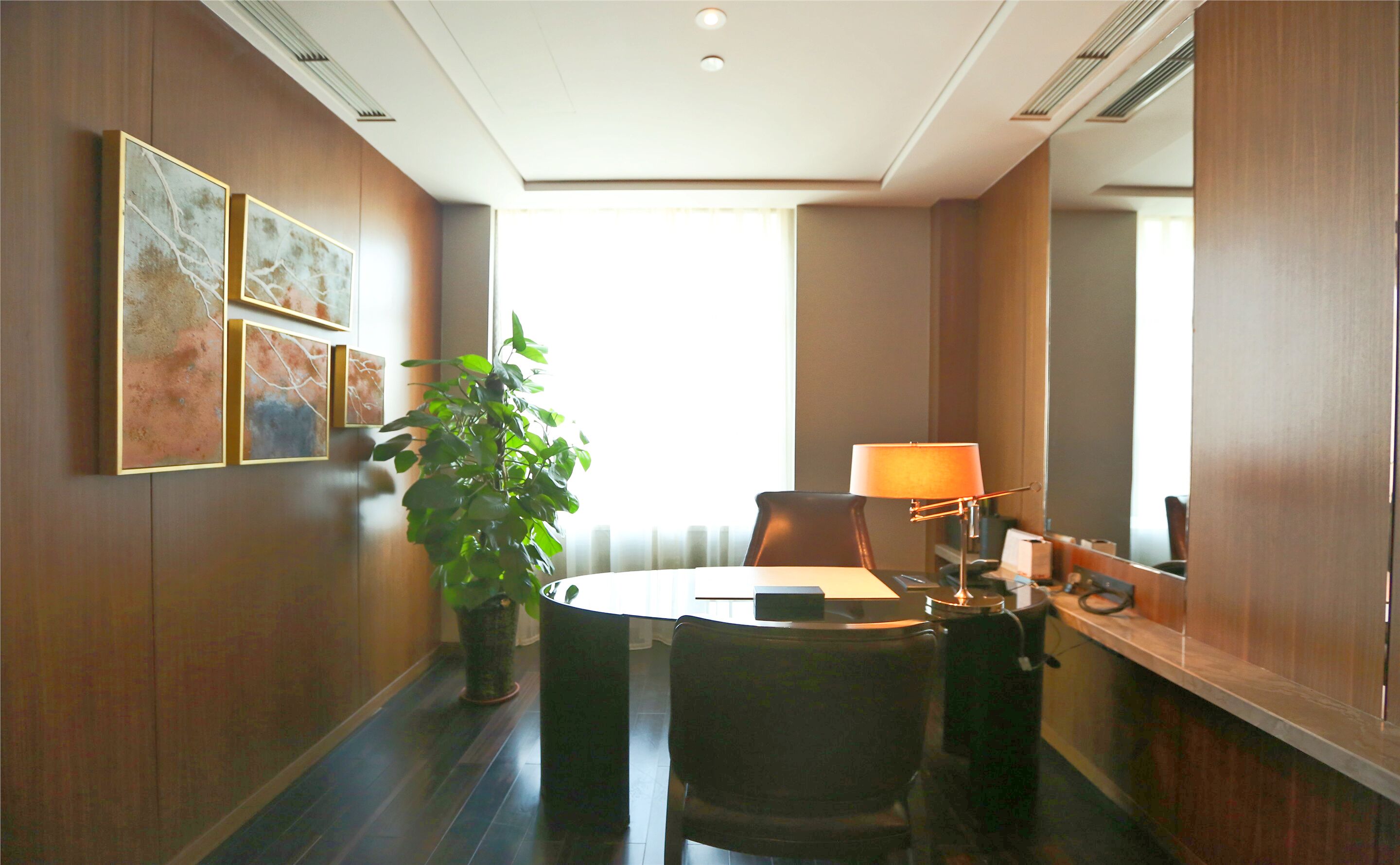 A hotel room with a dining table, chairs, a potted plant, a lamp, and artwork on the wall.