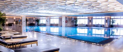 A modern indoor swimming pool with geometric ceiling design, lounge areas, and large windows.