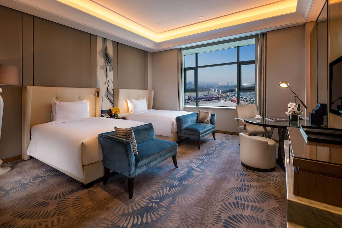 Una habitación de hotel con una cama grande, dos sillones, un escritorio y vistas a la ciudad a través de un amplio ventanal.