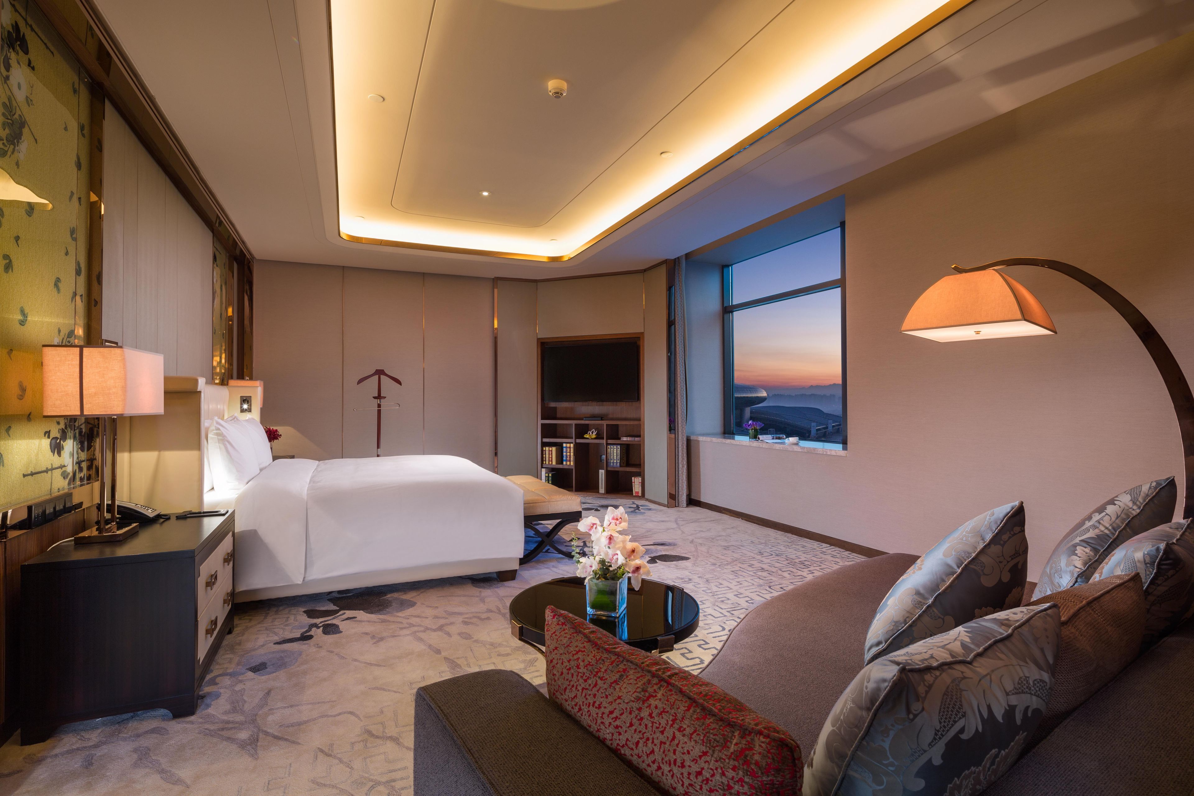A hotel room with a bed, a desk with a lamp, a sofa, a coffee table, and a large window with a view.