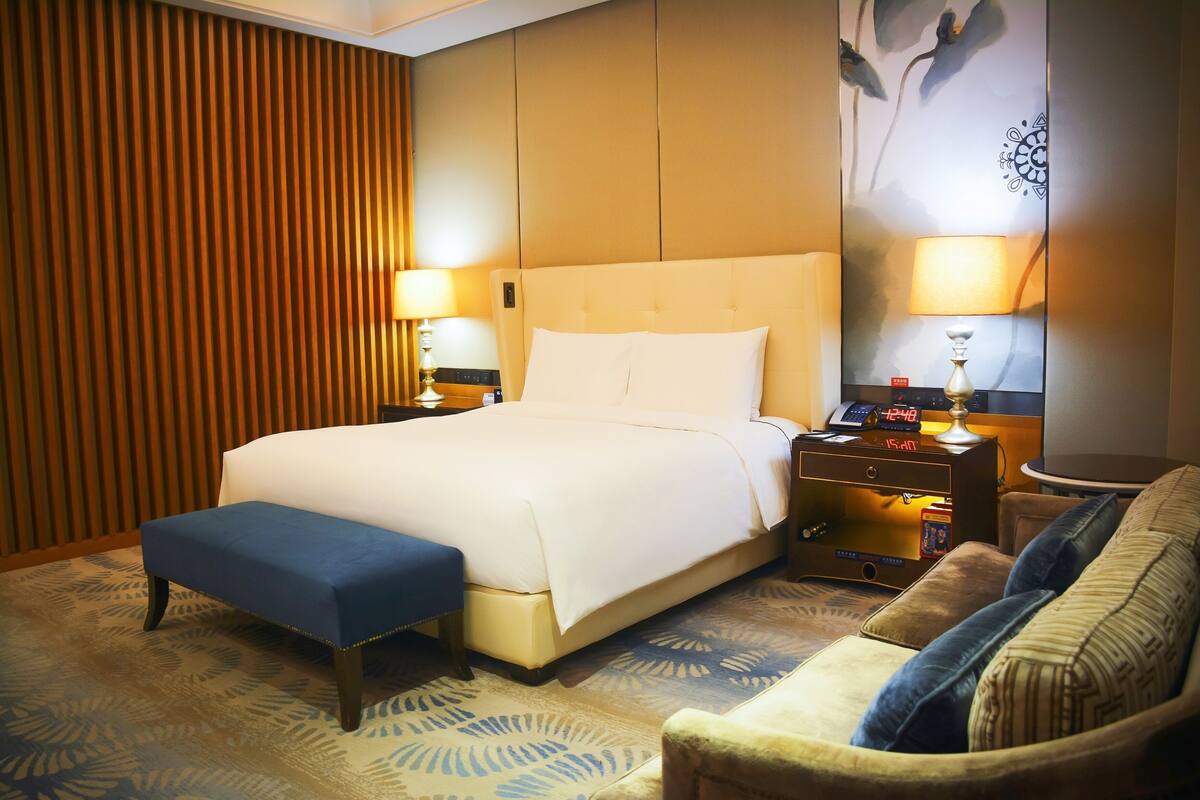 Una habitación de hotel con una cama grande, dos mesitas de noche con lámparas, un banco azul y un sofá.