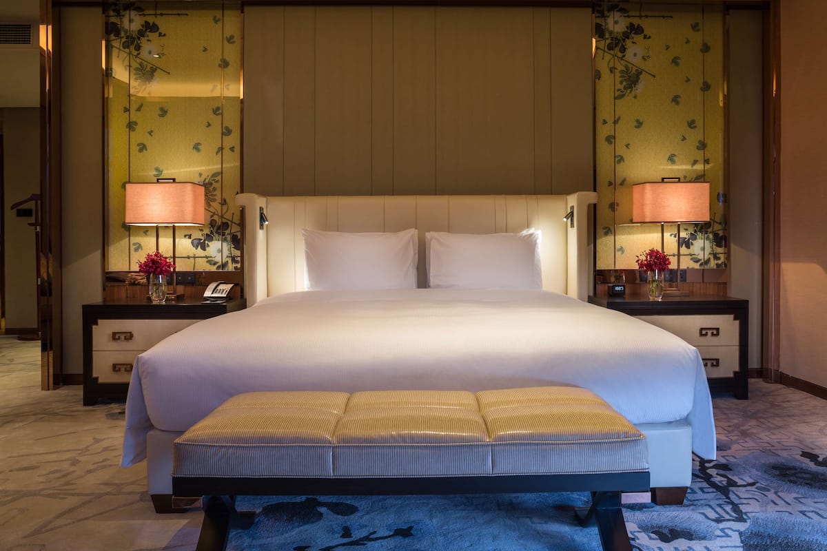 Habitación de hotel con una cama grande, dos mesitas de noche con lámparas, una mesita auxiliar, un banco y suelo alfombrado.