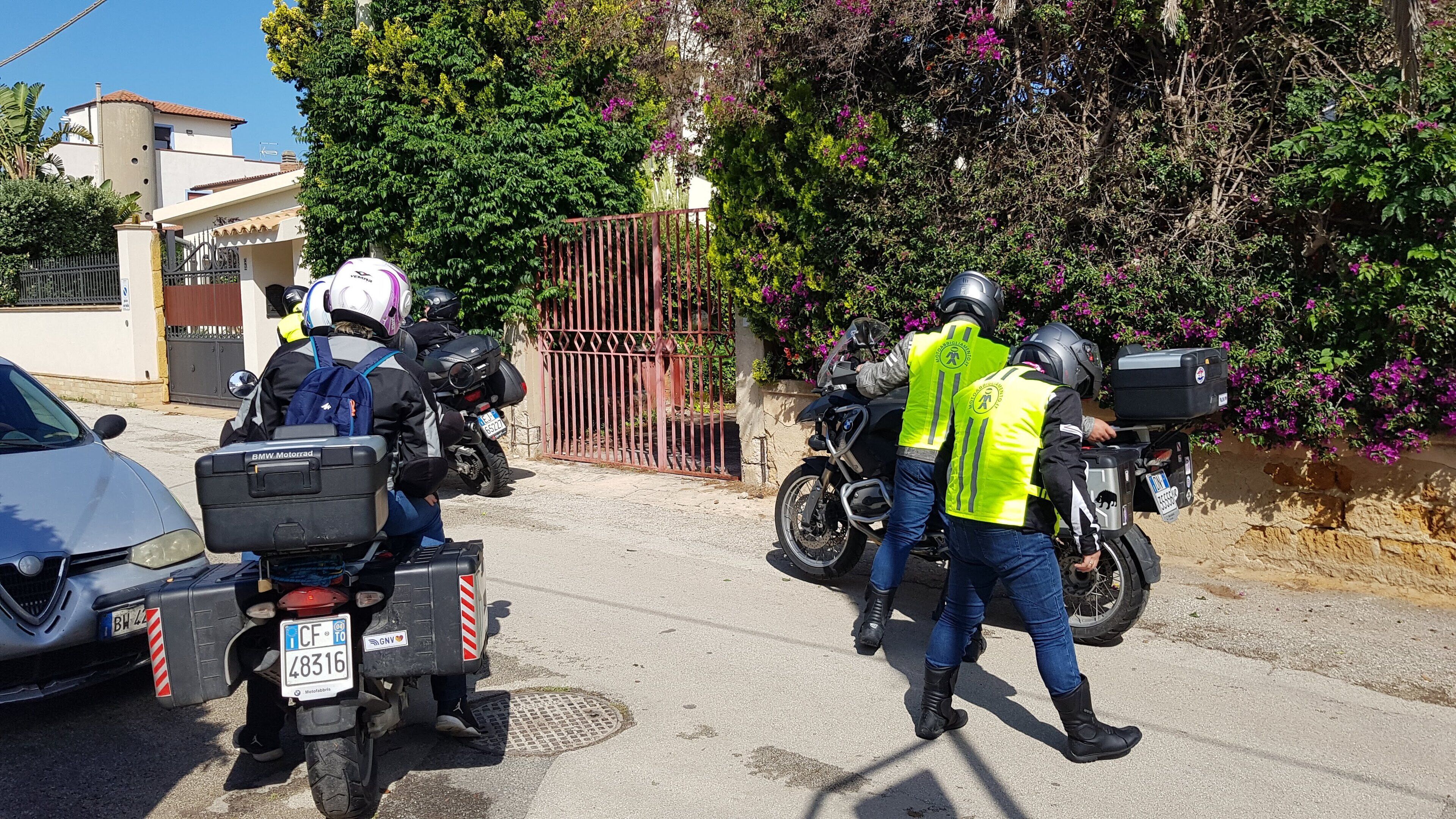Dos motociclistas con casco y chalecos reflectantes, uno con una mochila, estacionados en una calle con un coche y una reja al fondo.