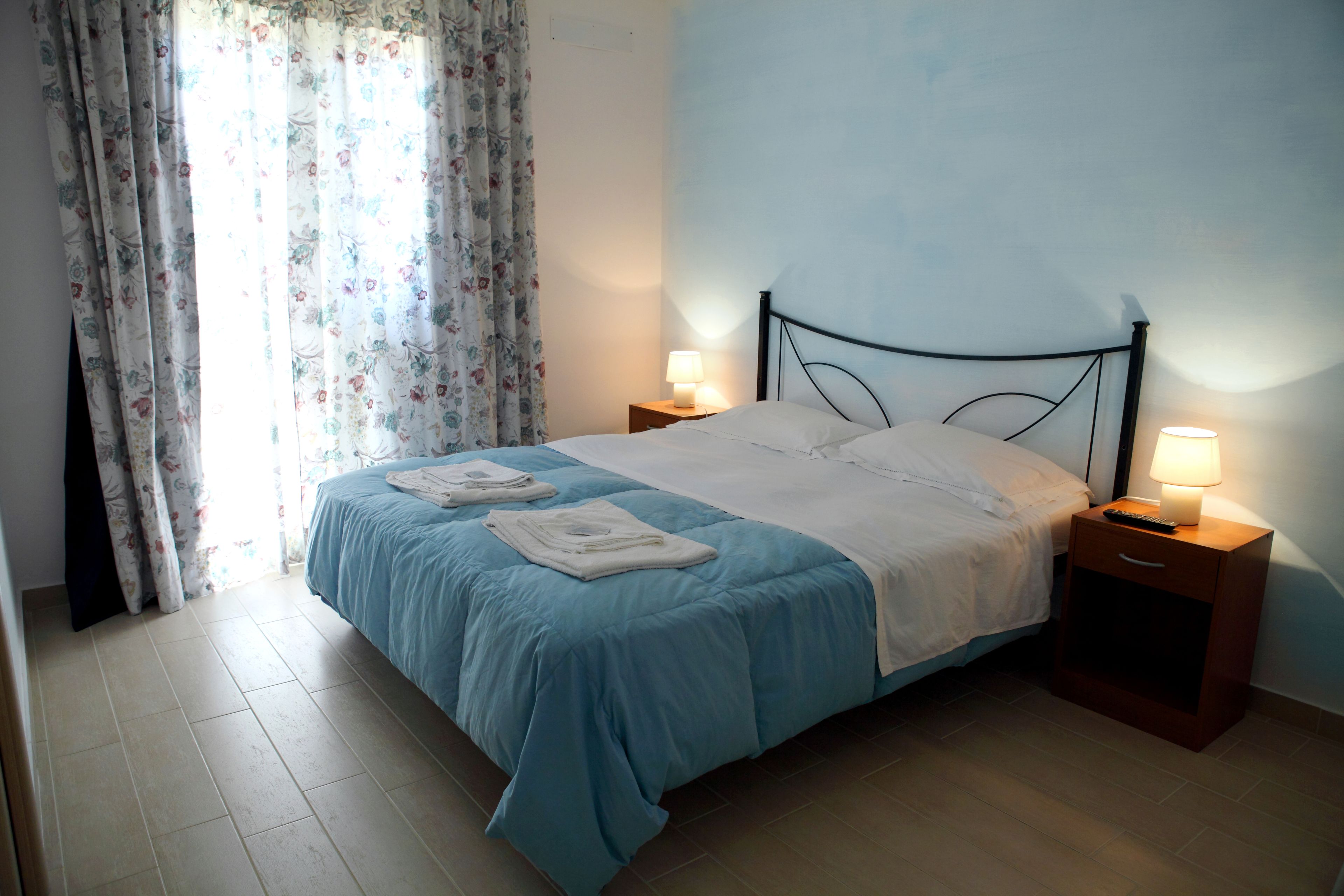 A neatly made bed with a blue and white duvet, two bedside tables with lamps, and a window with floral curtains.