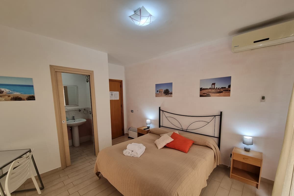 Een slaapkamer met een bed, nachtkastjes, een badkamer met een wastafel en een spiegel, en een wandairconditioner.