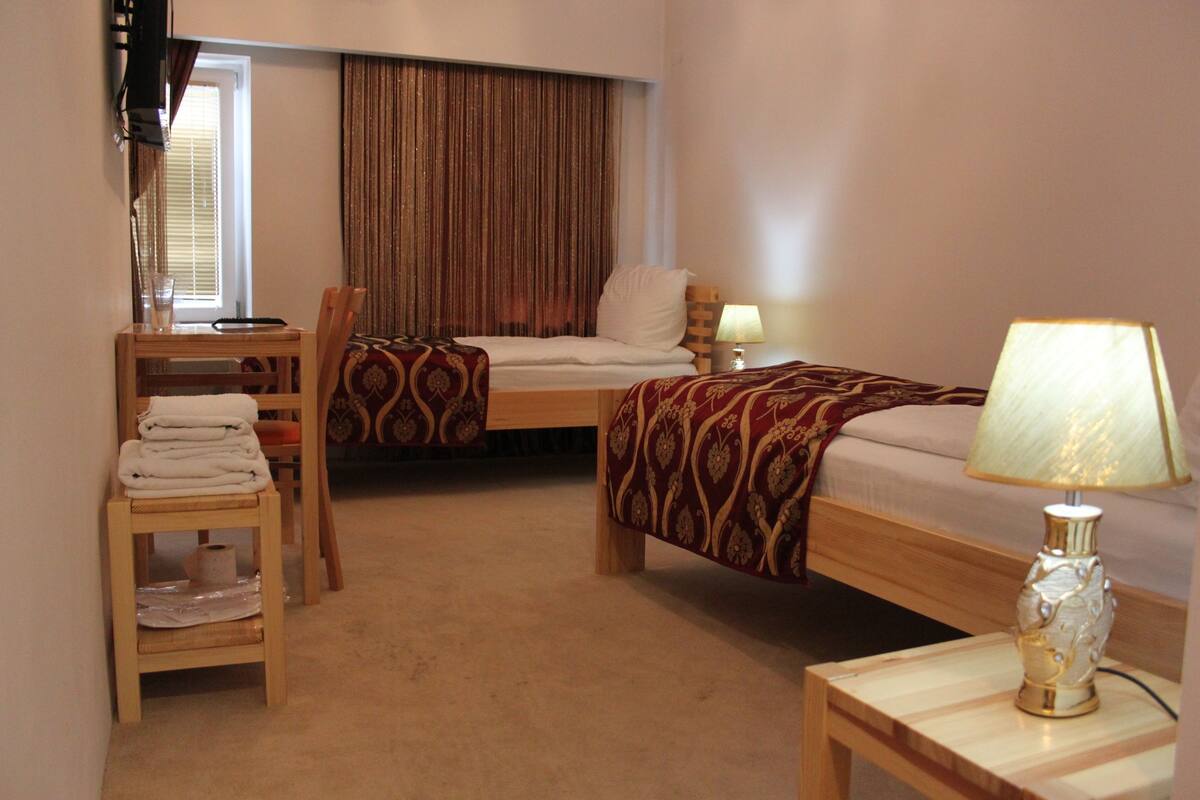 A hotel room with two single beds, a small wooden table, a chair, a bedside table with a lamp, and a window with curtains.