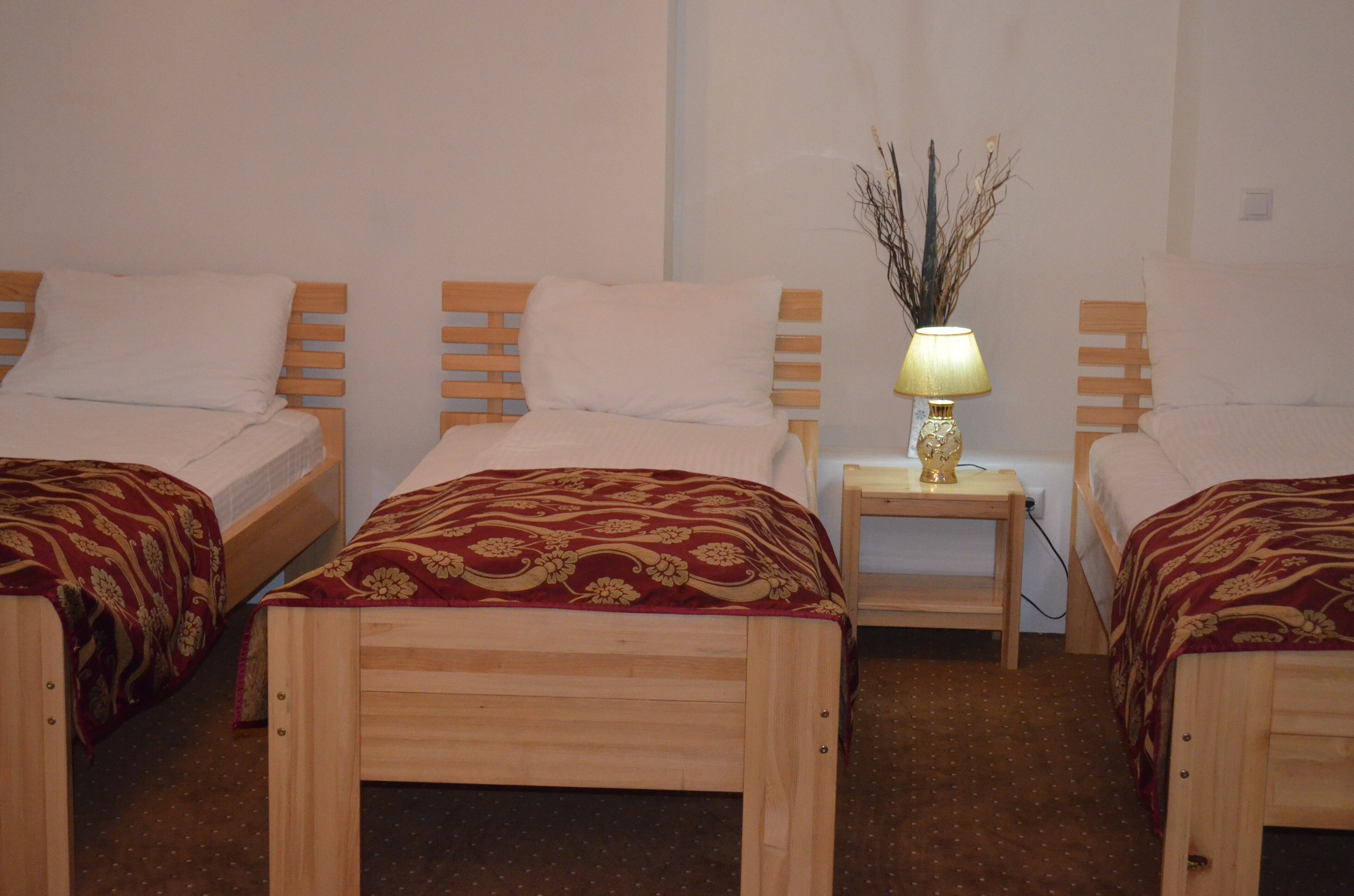 A hotel room with three wooden beds, each with a red and gold patterned bedspread. There is a small bedside table with a lamp and a decorative vase with branches.