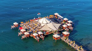 An aerial view of a floating island with multiple structures, including a central pavilion, sun loungers, and small huts, surrounded by clear blue water. - VONRESORT Golden Coast - All Inclusive (Manavgat)