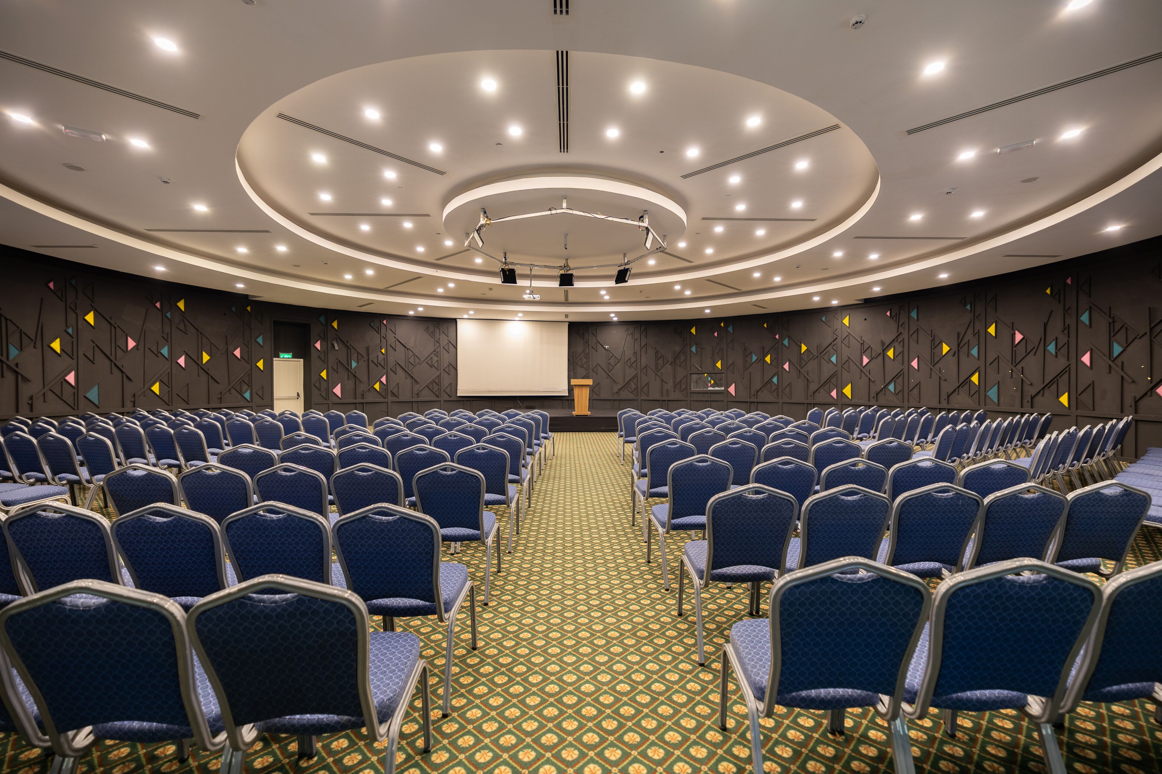 A conference room with rows of blue chairs, a patterned carpet, and a large screen at the front.