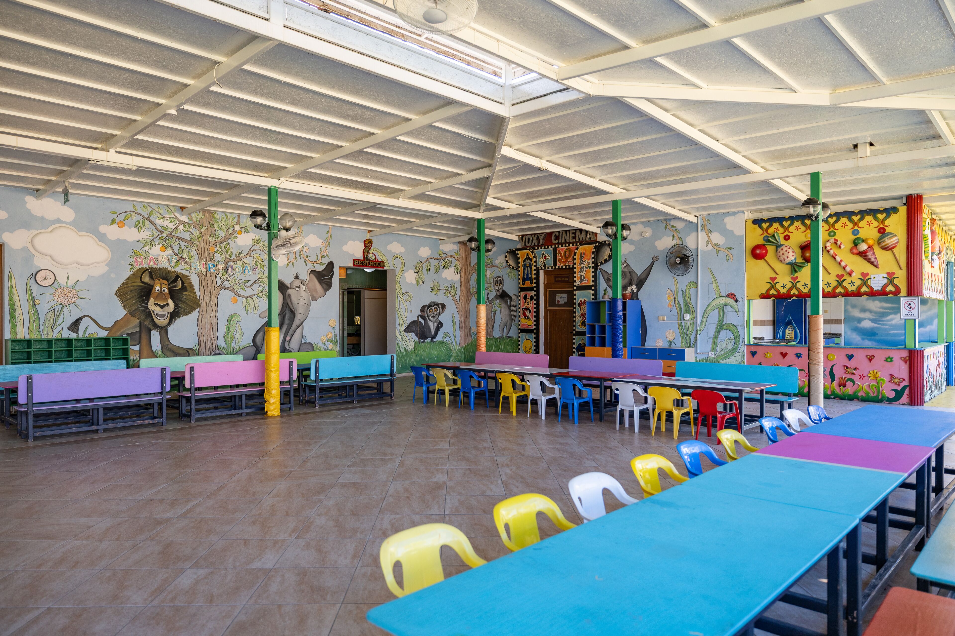 Un área de juegos para niños con mesas y sillas de colores, murales en las paredes y un techo con diseño de cuadrícula.