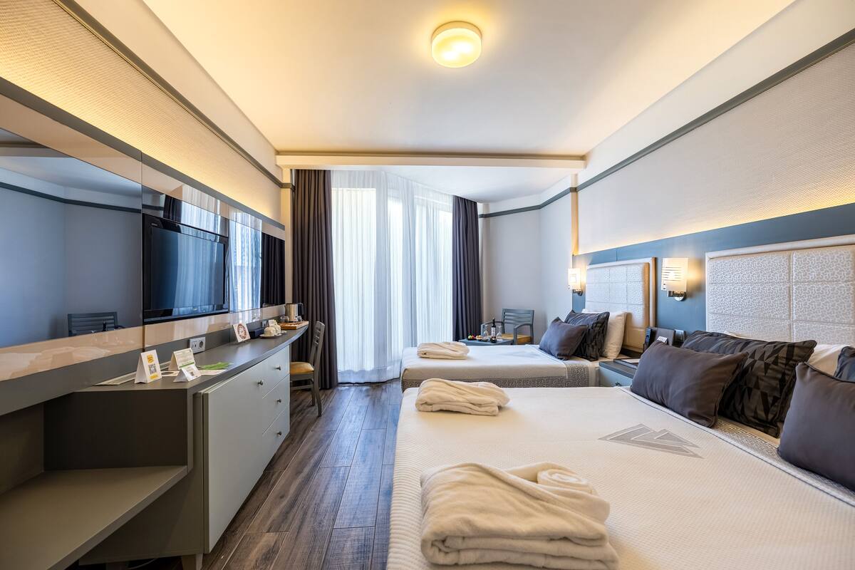 A hotel room with a bed, bedside tables, a desk, a TV, and a window with curtains.