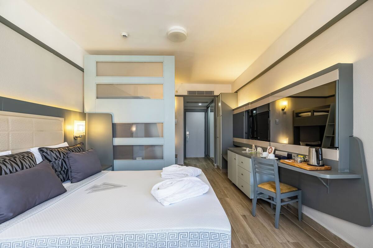 A hotel room with a large bed, a desk with a chair, a mirror, and a kitchenette.