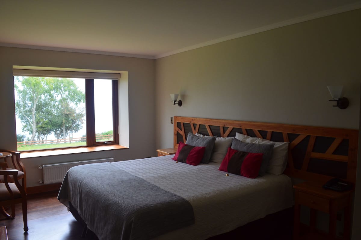 Quarto com cama grande, janela vista para árvores, cabeceira de madeira e mesa de cabeceira com abajur.