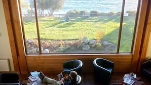 Una habitación con un ventanal grande que da a un área verde y vista al agua. Hay dos sillones negros y una mesita con un libro.