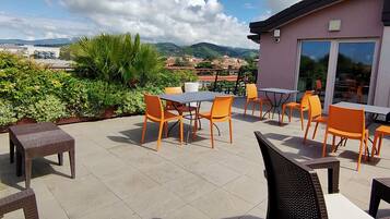 Un patio exterior con mobiliario de mimbre y sillas anaranjadas, con vistas a un paisaje urbano y colinas.